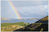Rainbow hitting island in loch surrounded by hills.