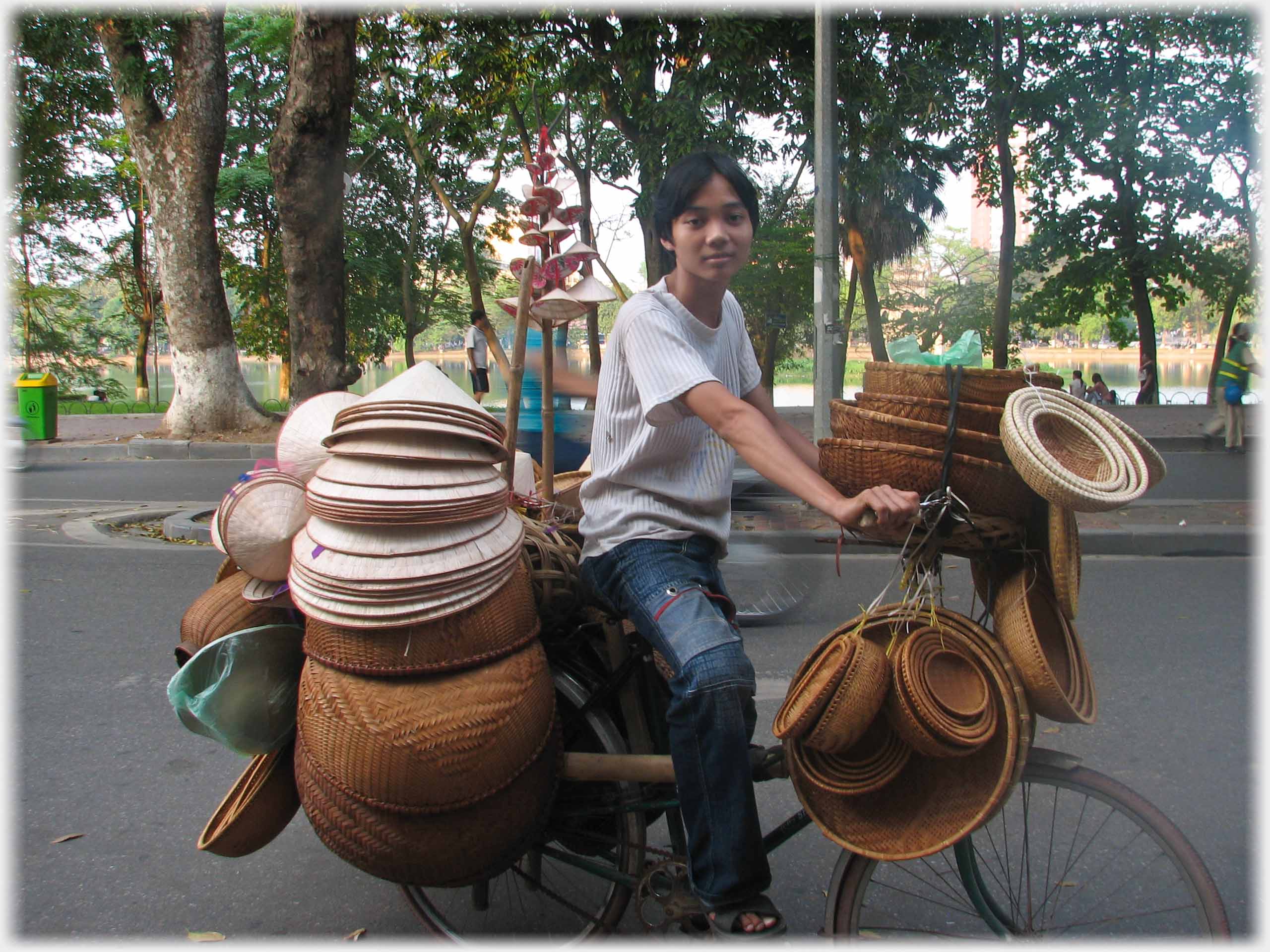 Young vendor of indeterminate sex on bike loaded with hats and baskets.