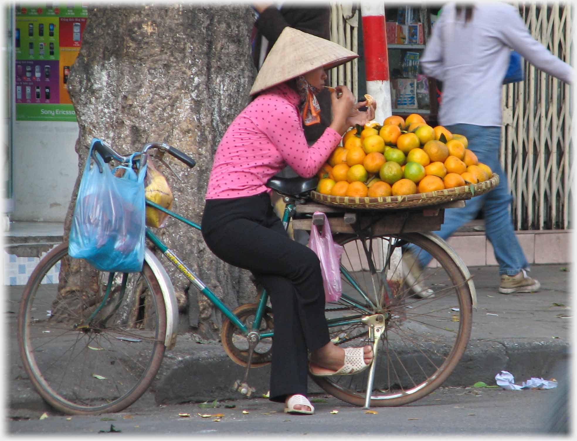 Woman sitting on bar of bike with pile of oranges, apparently eating.