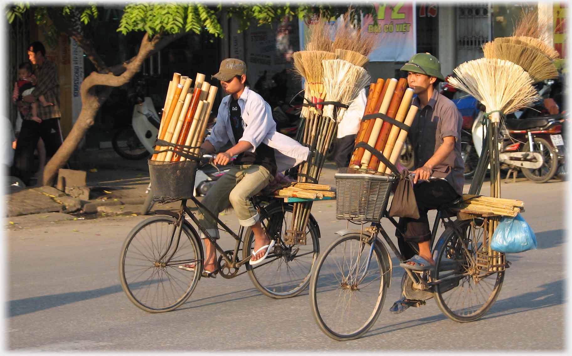 Two vendors cycling together with brushes behind and bamboo pipes in front.