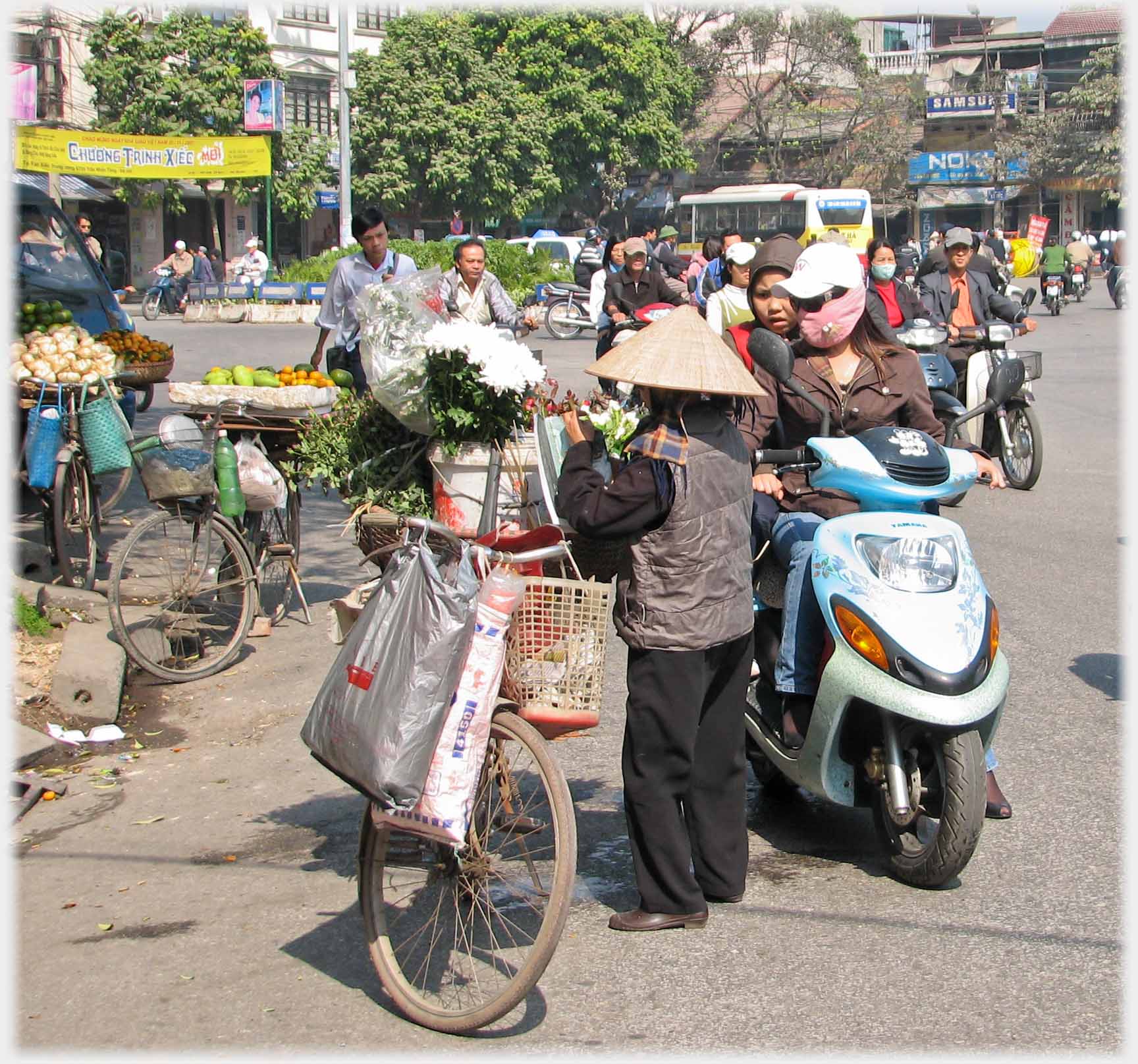 Woman by her bike on which are buckets of flowers, woman on scooter stopping to look.