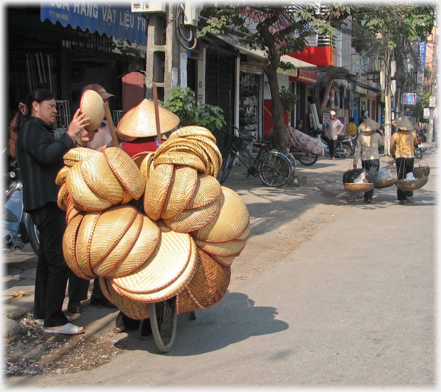 Bicycle loaded with baskets, woman examines one of them, a conical hat on a red shirt peeks above.