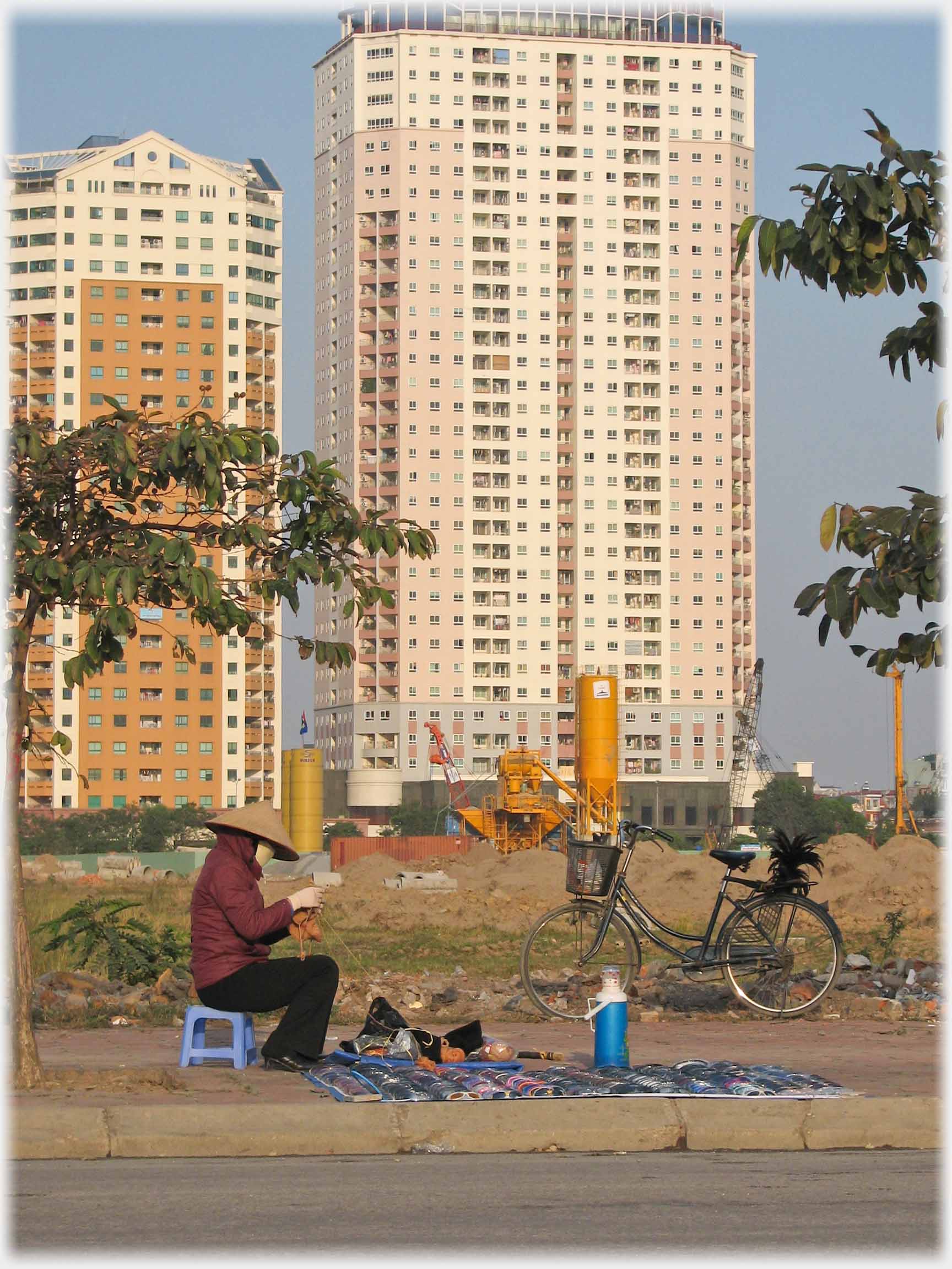 Two thirty story tower blocks form background to a woman sitting on the pavement knitting with her large selection of sunglasses laid out and a bike nearby.