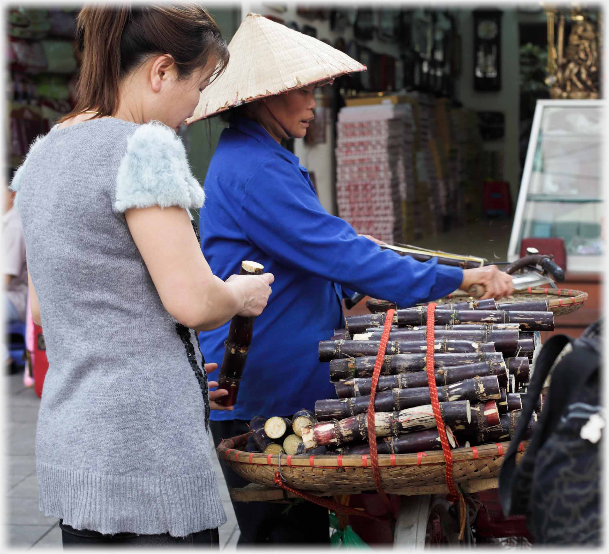 Woman in hat with knife by piles of sugar cane, customer holding one piece.