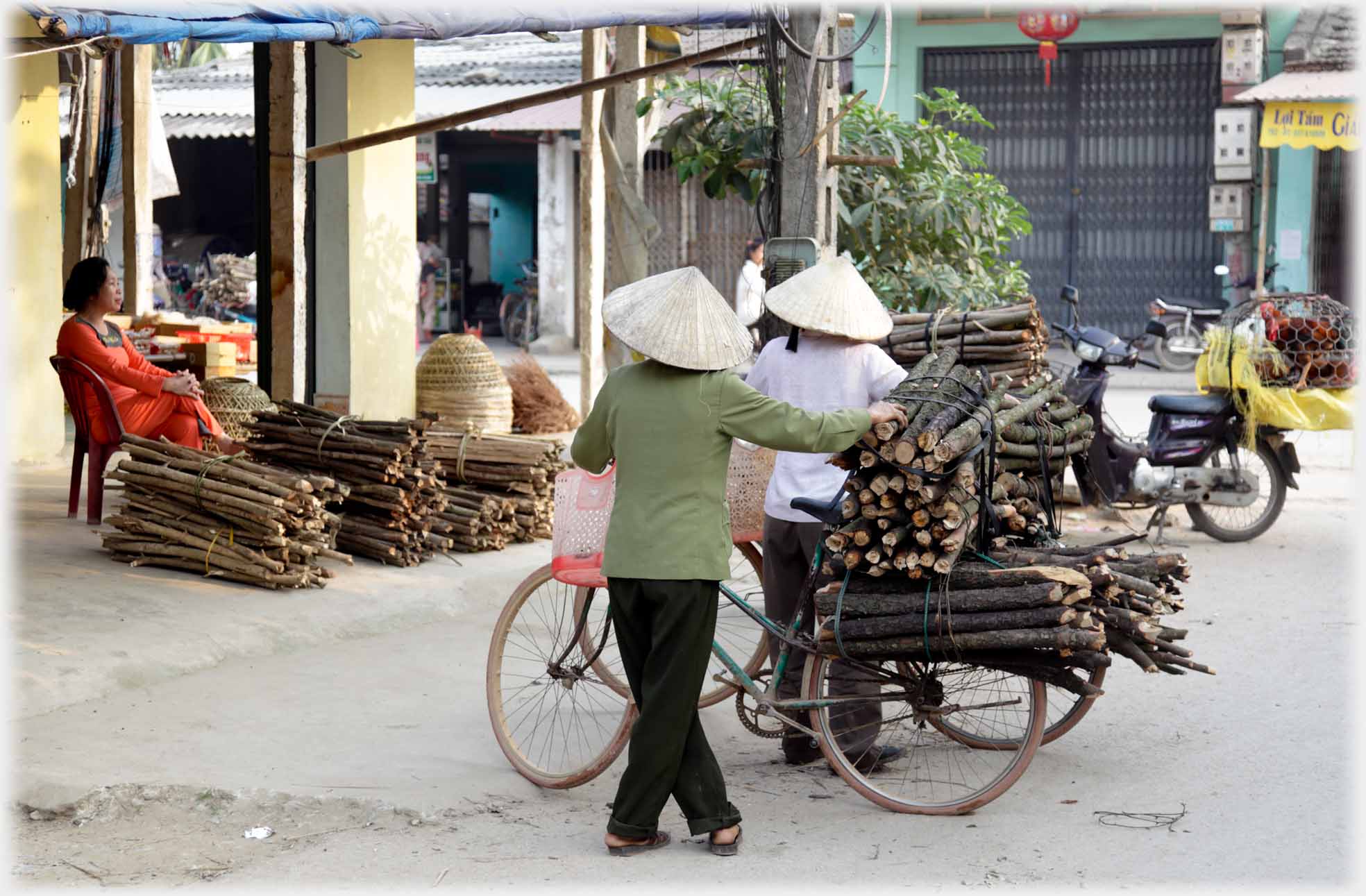 Two women with bikes piled with faggots, one woman sitting by bigger bundels of faggots nearby.