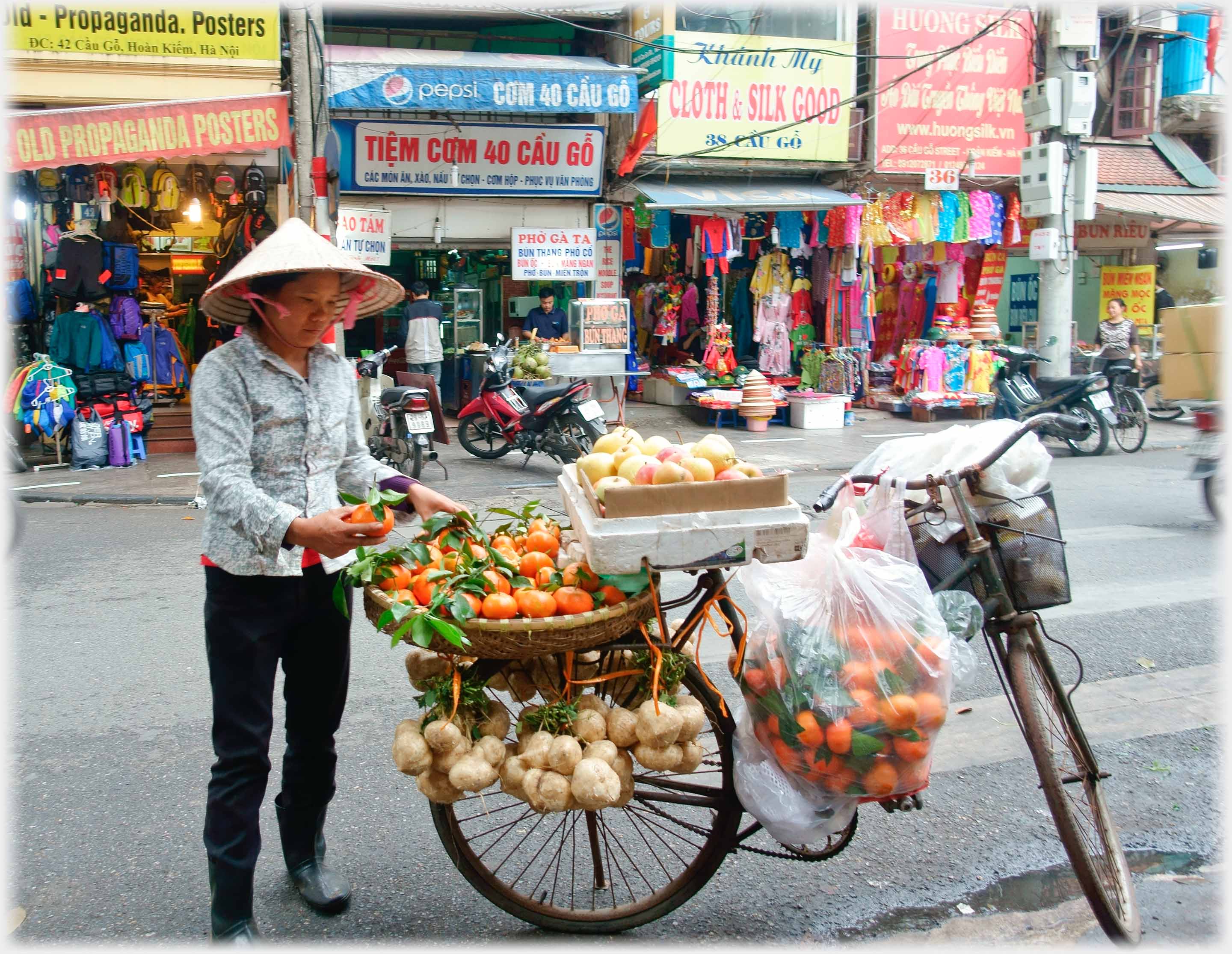 Woman standing beside a bicycle loaded with oranges, apples and vegetables.