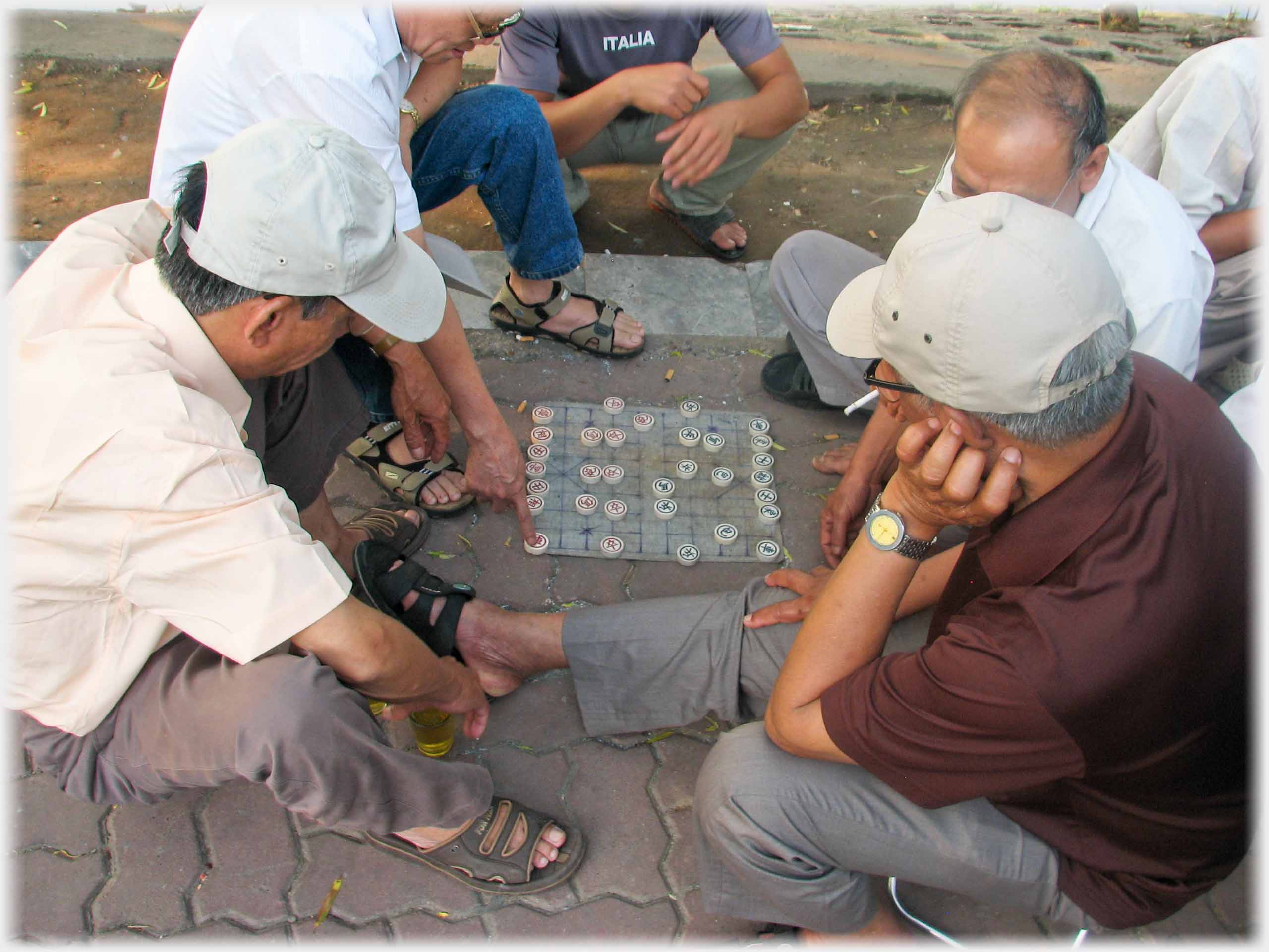 Group around cloth 'board', a finger on a piece; cigarette and tea apparent.