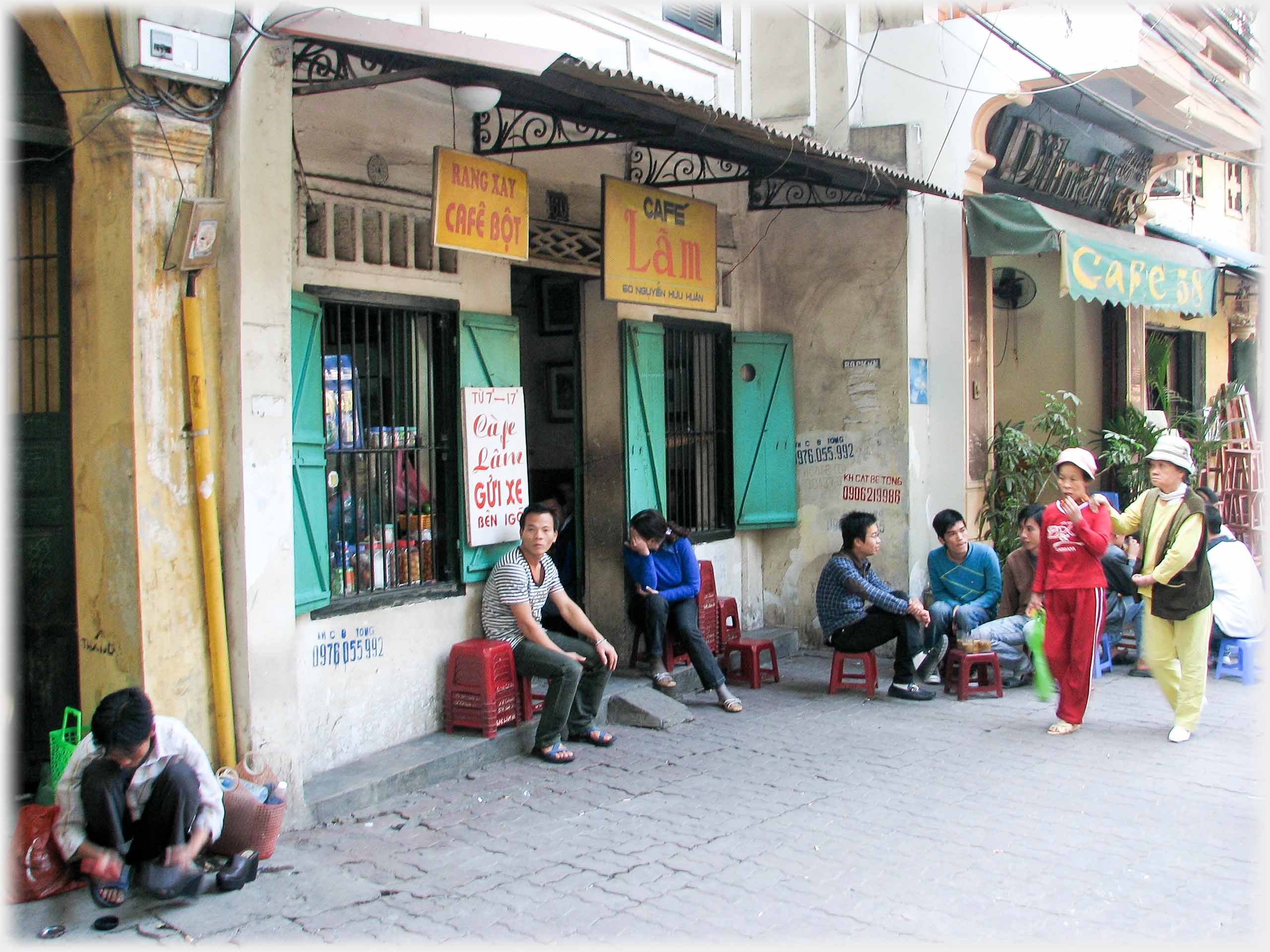 Pavement in front of Cafe Lam. Various people sitting. Woman leaning on another walking past. Man cleaning shoes.