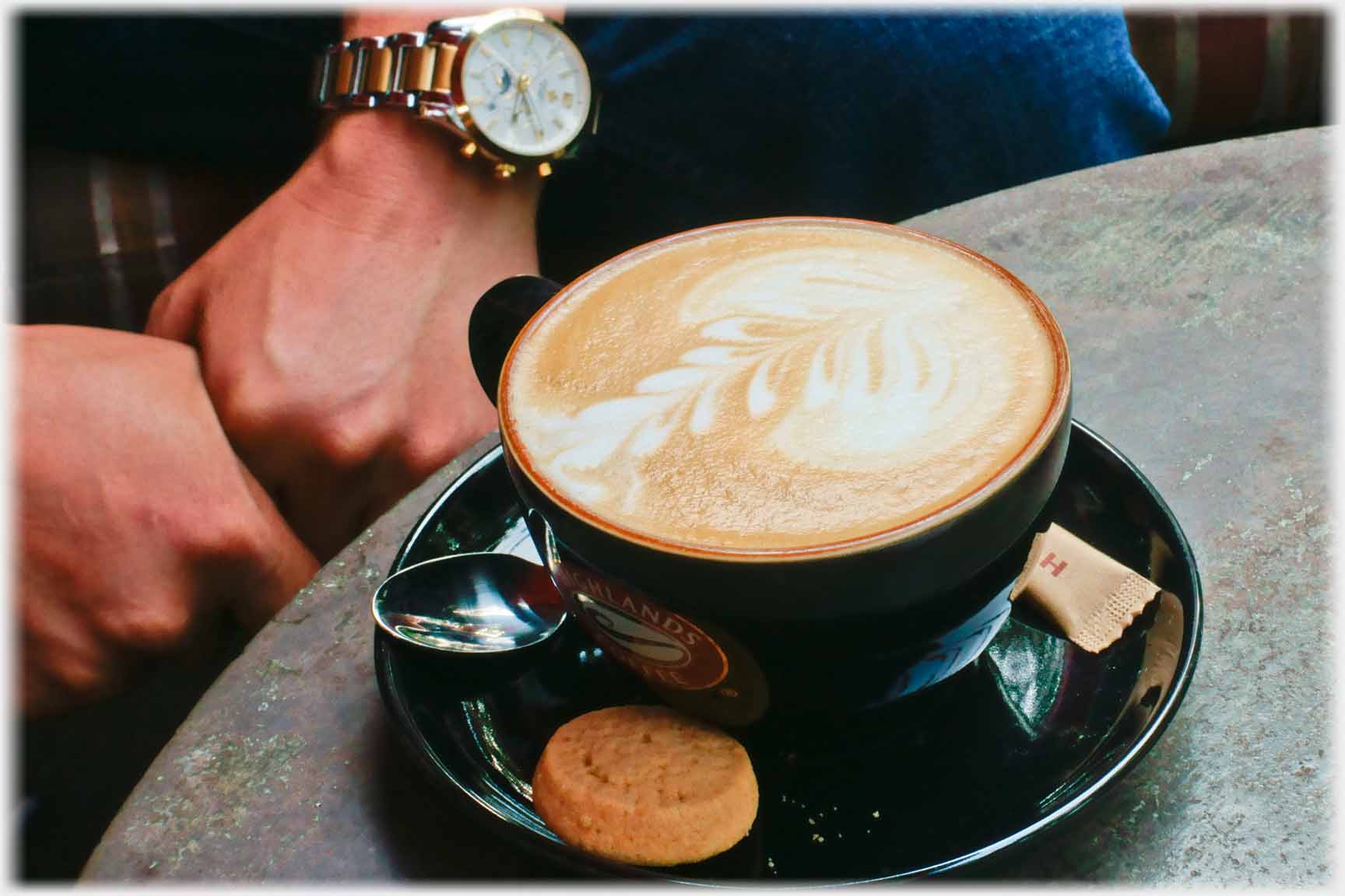 Black western styly coffee cup with Highland badge on side and a leaf configured in foam. Hands and watch beside it.