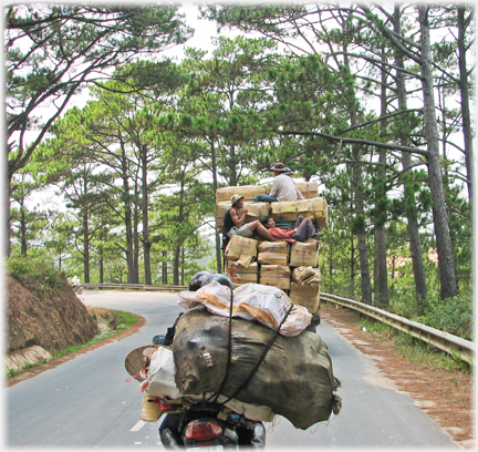 Load obscuring rider falling off bike, in front of it truck laden high with boxes, three men on these.