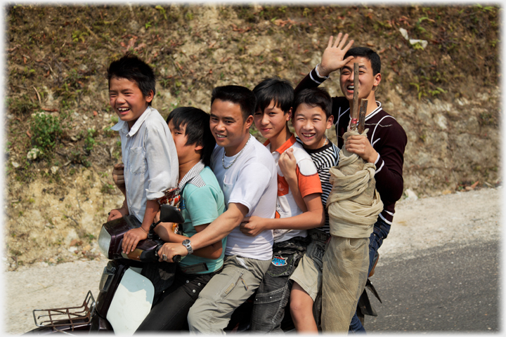 Smiling man driving, another man holding nets for fishing and waving, and four teenage boys on a bike.