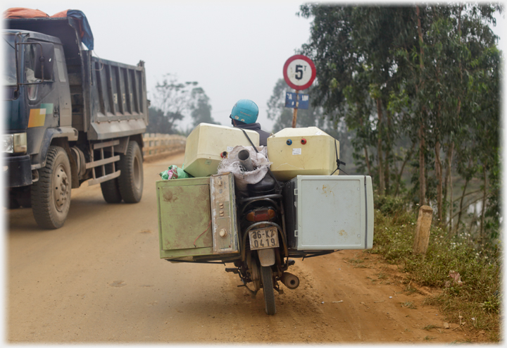 Motorbike with machines strapped to either sidde and on top, lorry on other side of road, 5 ton weight limit.