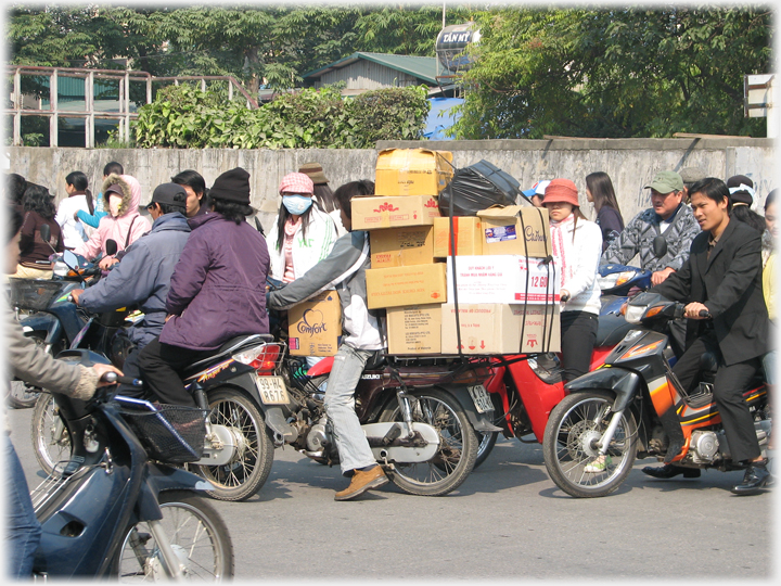 Motorbike laden to head height with boxes apparently trapped amongst a crowd of motorbikes.