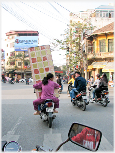 Woman pillion holding bed, man looking across at her from next bike.