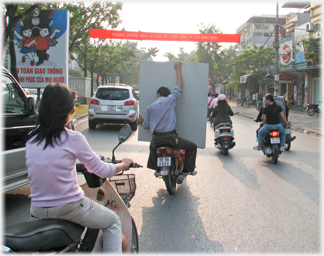 In traffic with woman near, man holding board centre and woman with suitcase beyond.