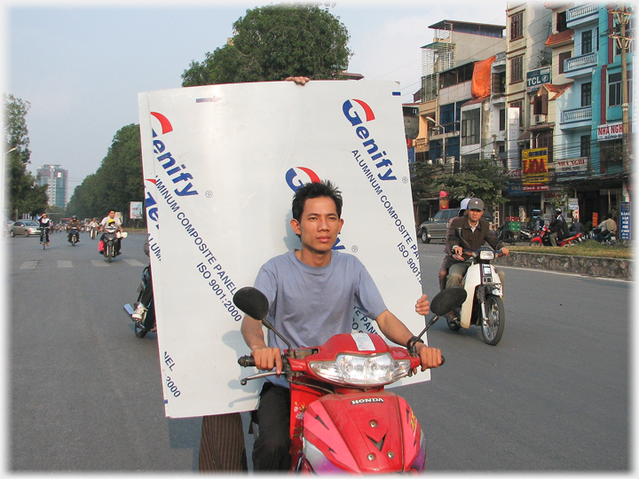 Man driving red Honda with large panel behind him, held by two sets of fingers.