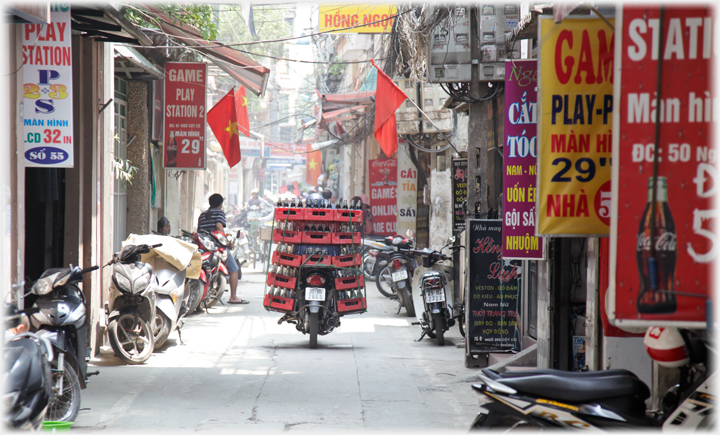 Motorbike in motion down lane with thirteen crates of Coca-cola stacked on it.