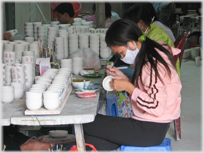Girl sitting painting pot, many stacked on tables beside her.