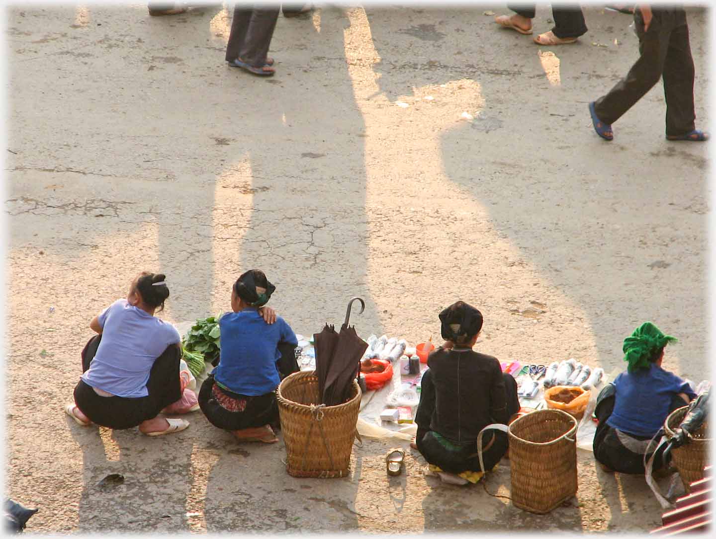Four women sitting at the roadside with goods laid out in front of them, seen from above and behind.