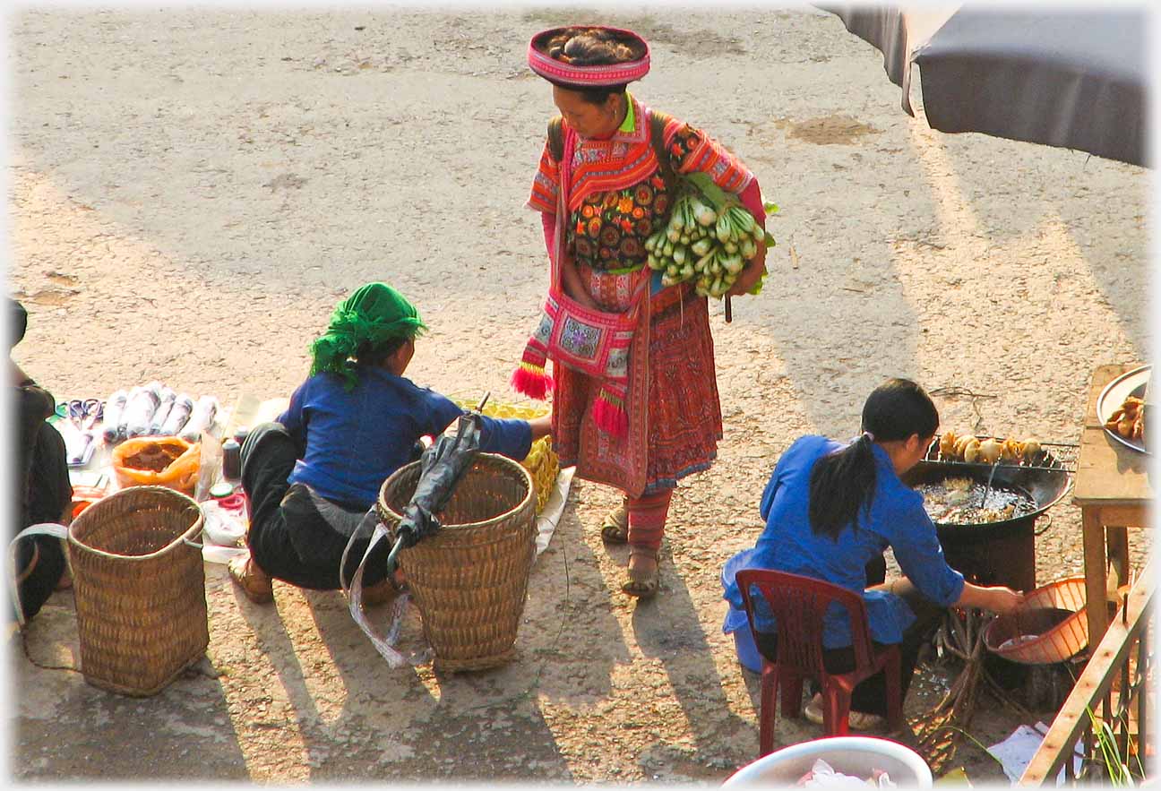 Customer standing with bundle of vegetables by baskets and woman sitting cooking.
