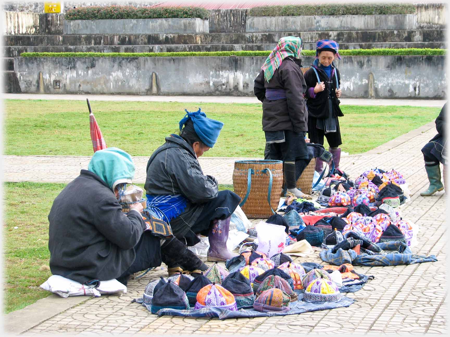 Piles of hats in front of women on a paved area with grass around.