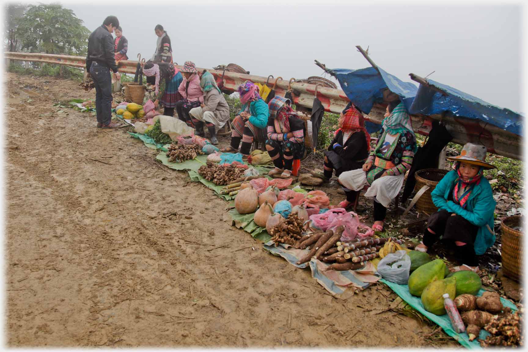 Line of produce on mats in front of line of women leaning against barrier.