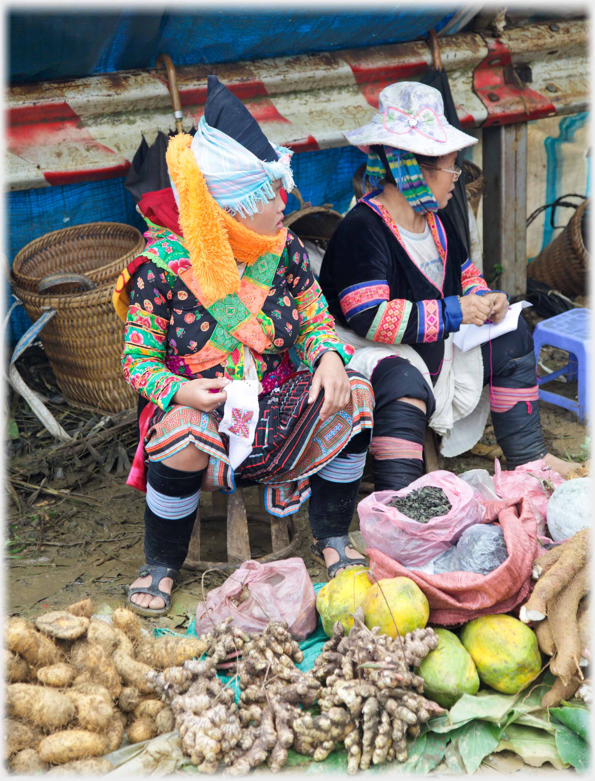 Two women sitting on stools behind their vegetables wrapped in a variety of ethnic clothes.