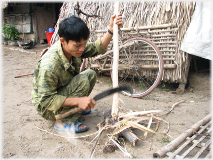 Man crouched down using a matchet to shape a pole.