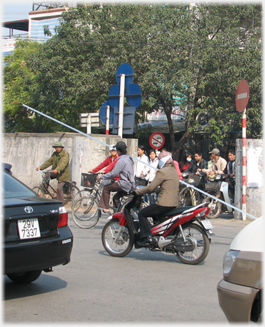 Cars and motorbike with cyclists, one of whom has a very long pole on his shoulder.