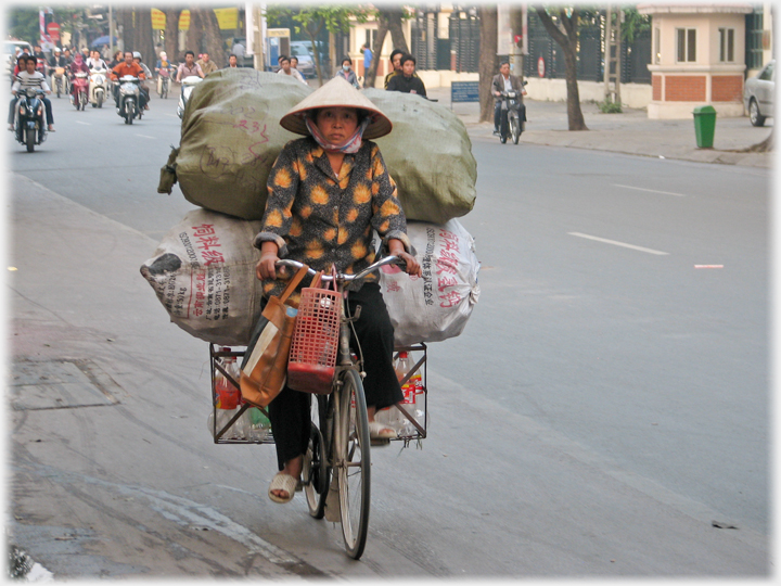Woman with hat and two large sacks on the back of her bike as she cycles down a Ha Noi street.
