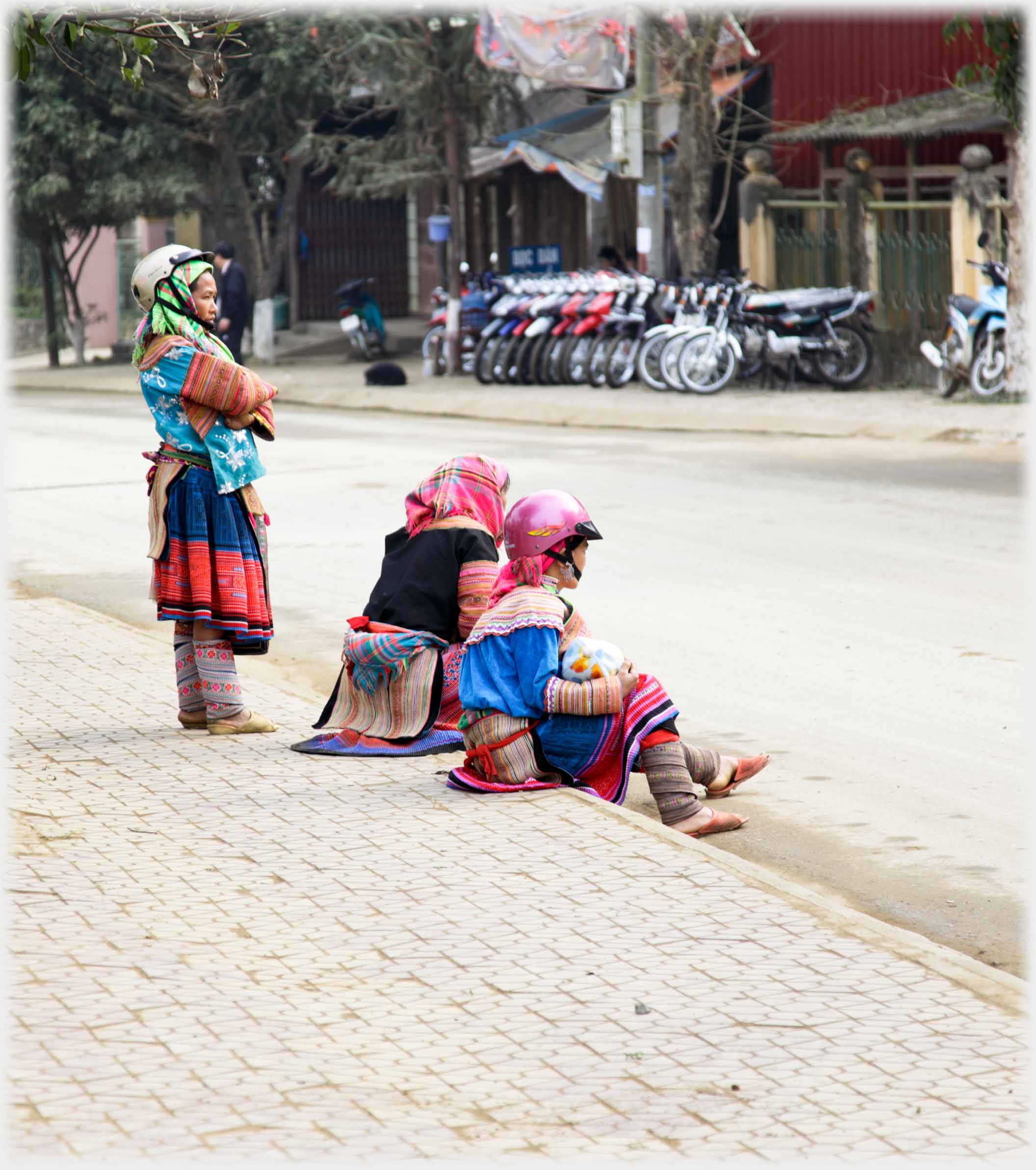 Hmong woman standing with crash helmet over head scarf, similar sitting on kerb next to woman in headscarf. Motorbikes for sale in background.