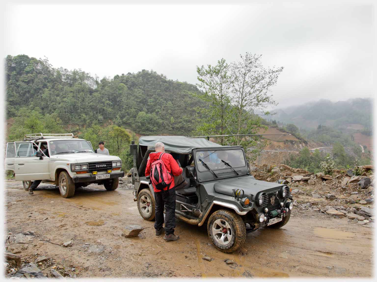 Man standing beside Jeep, car behind, mud road.