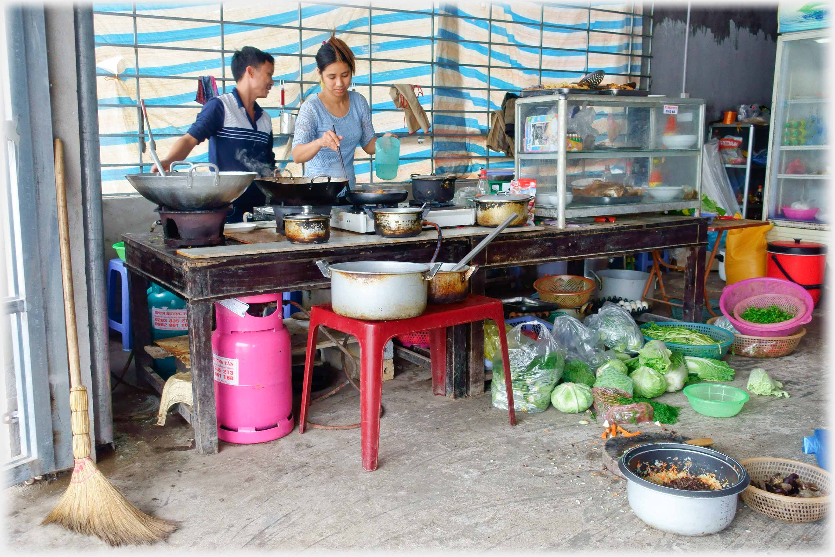Man and woman behind table with cooking utensils on top, many vegetables and pots on the ground, and other general clutter.