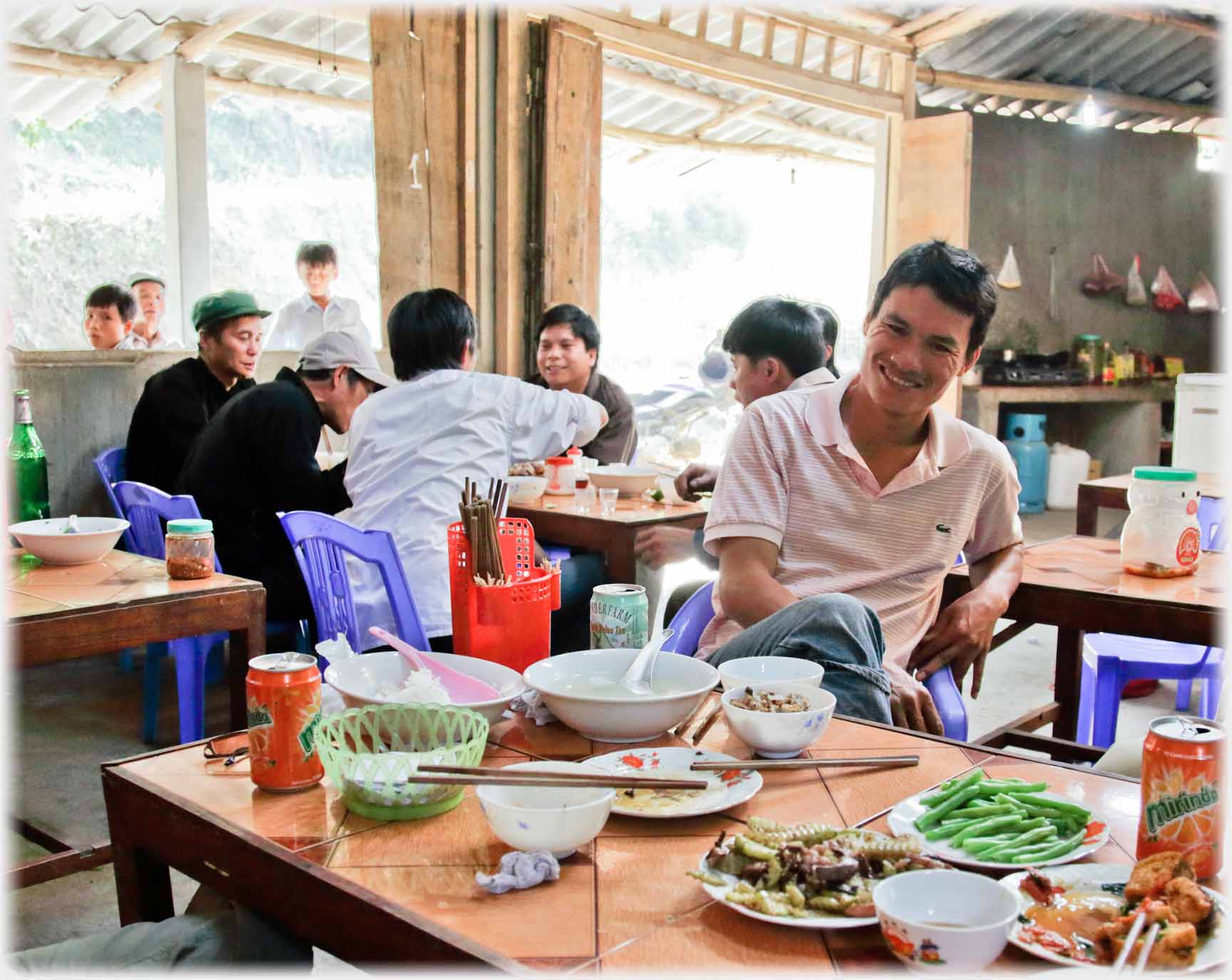 Man sitting smiling at table with remains of meal, other tables beyond.