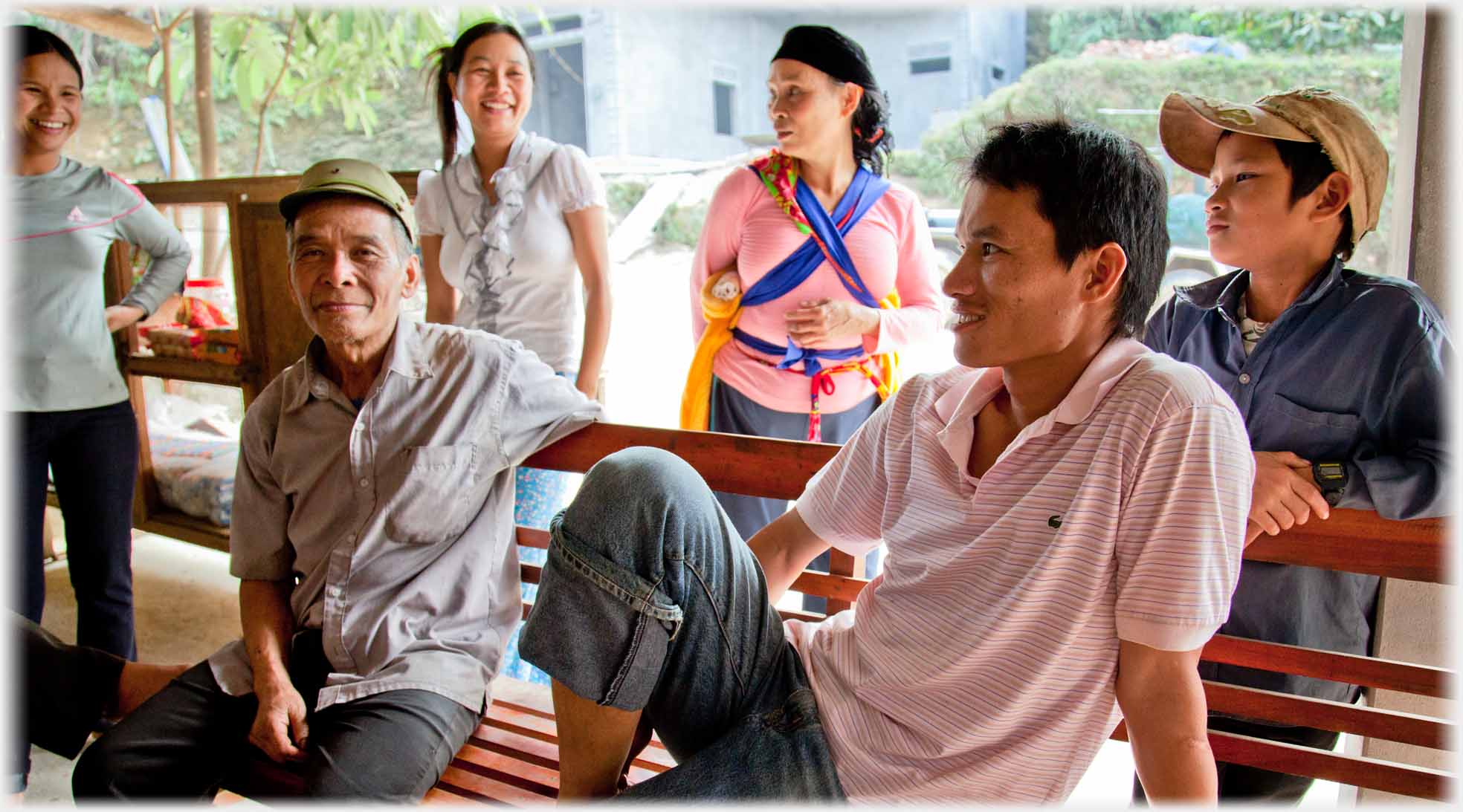 Two men on bench, younger with leg up, three women and boy behind.