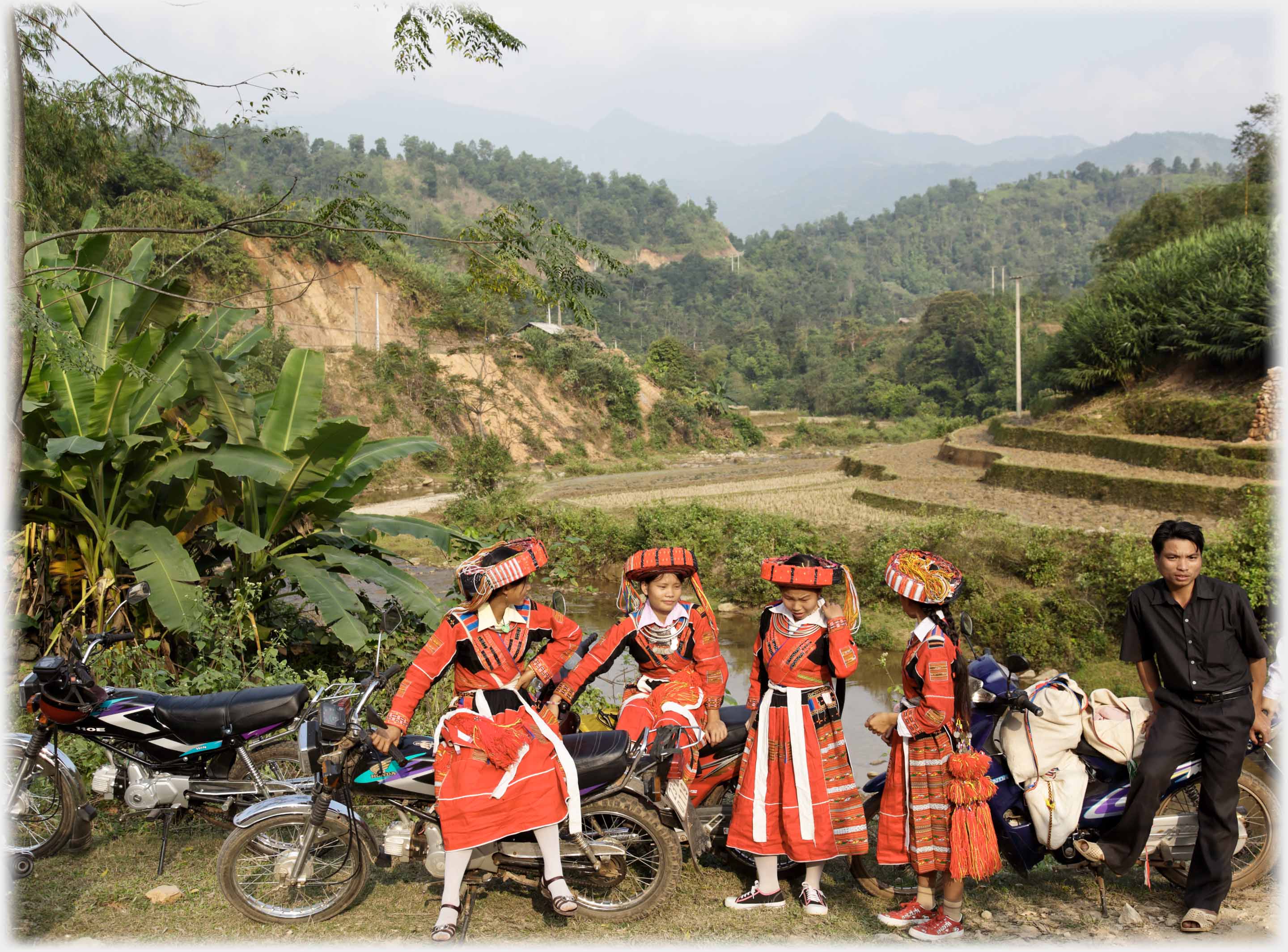 Four women in bright red clothes by motorbikes, man, valley beyond.
