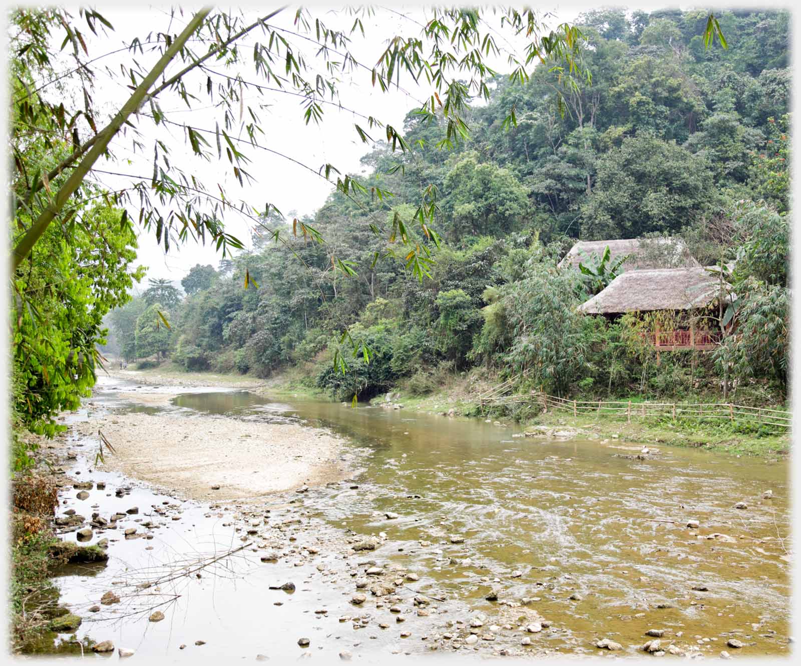 Shallow river across which two thatched huts amongst woods.