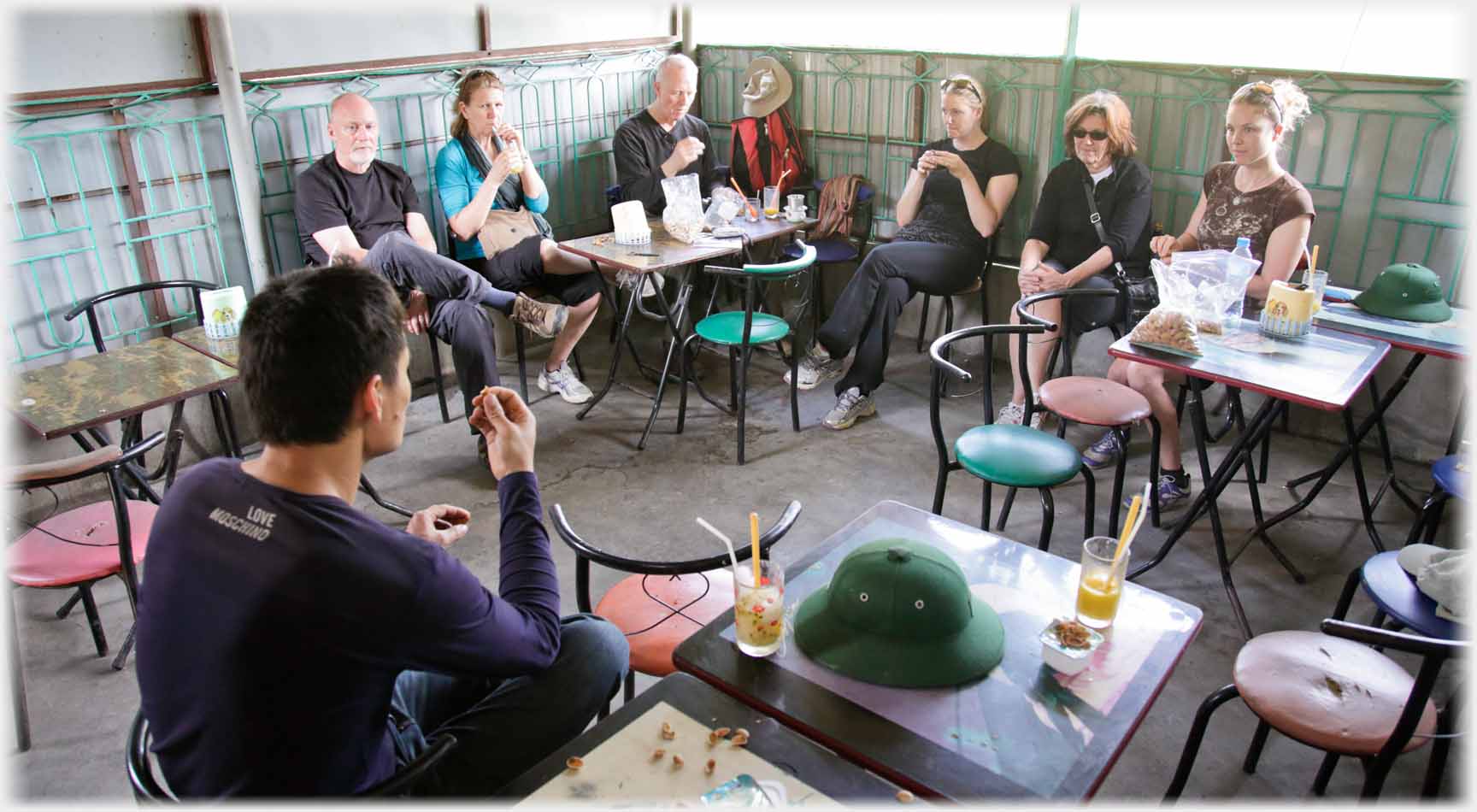 Man back to camera, six westerners along two walls, utilitarian furniture.
