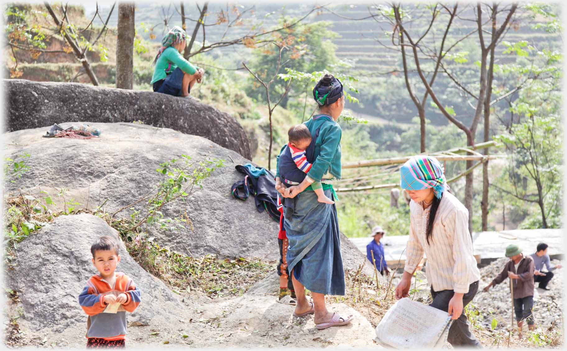 Woman with baby on back watching workers, child watching camera.