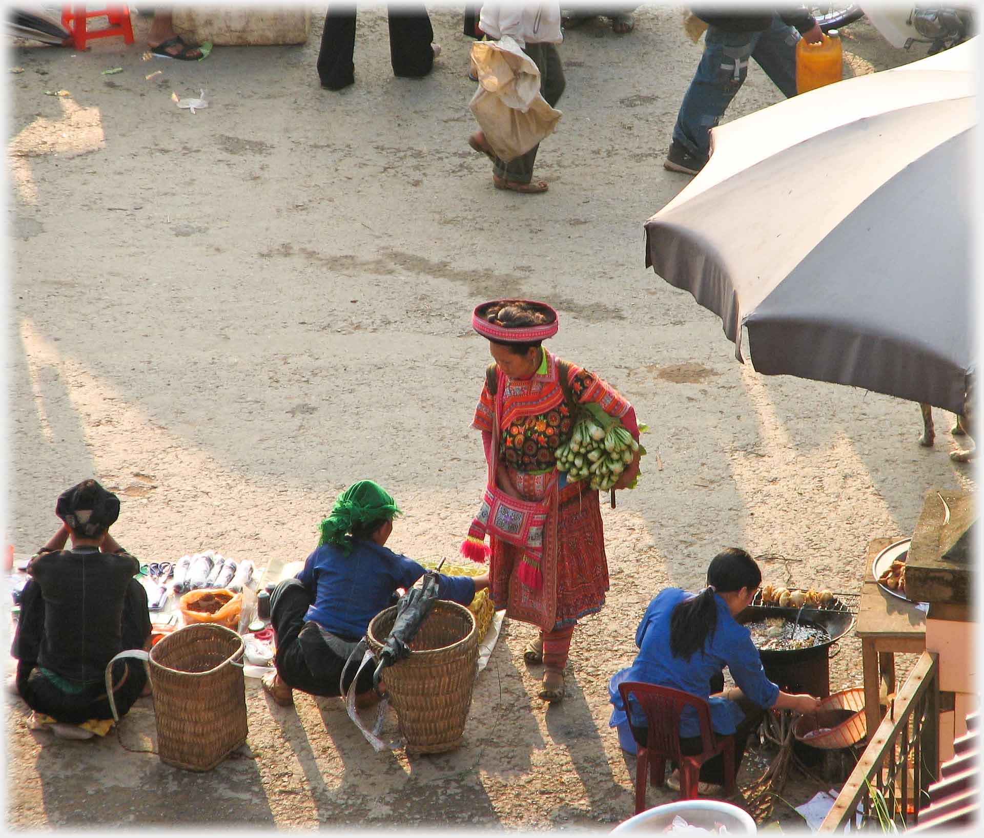 Woman standing with bundle of veg under her arm, talking to seller on the ground.