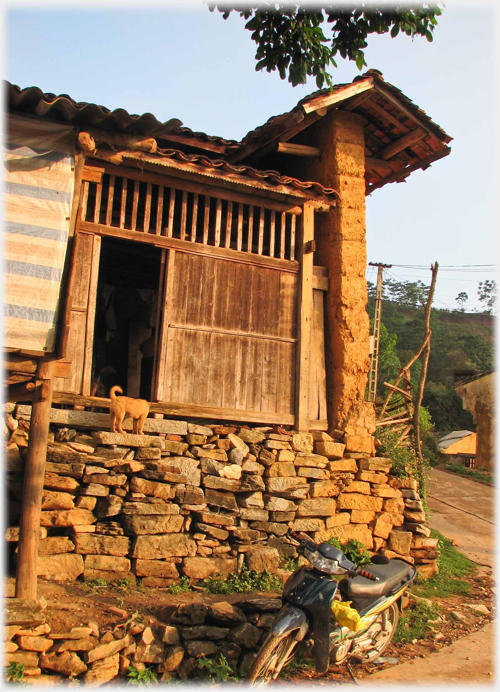 Wooden entrance to corner houose with un-mortared  stones below and angled roof over mud wall.