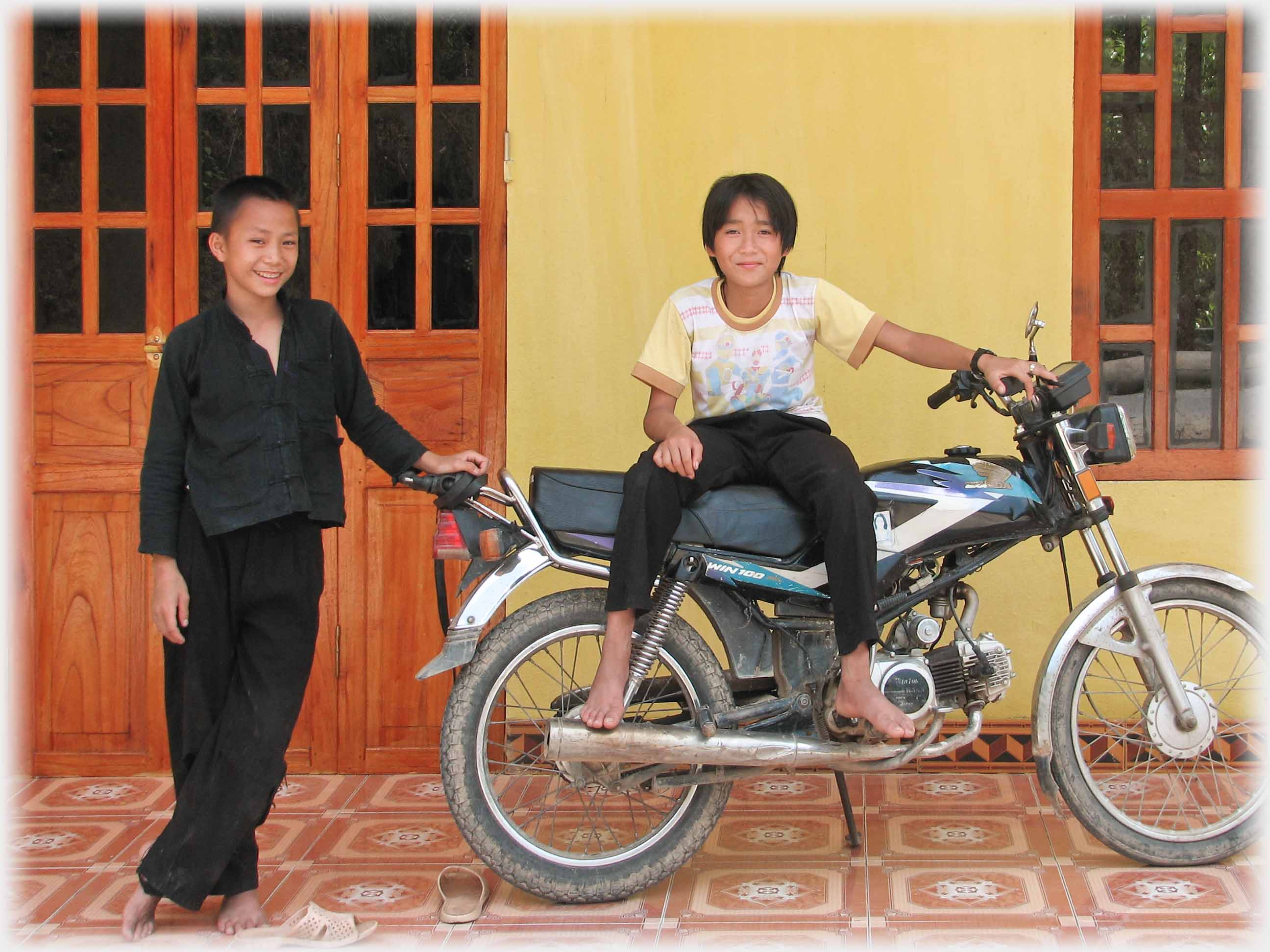 Youth on motorbike, another next to him, yellow wall behind.