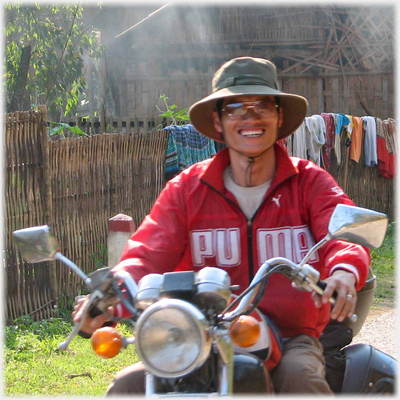 Han in red jacket and wide hat smiling broadly on motorbike.
