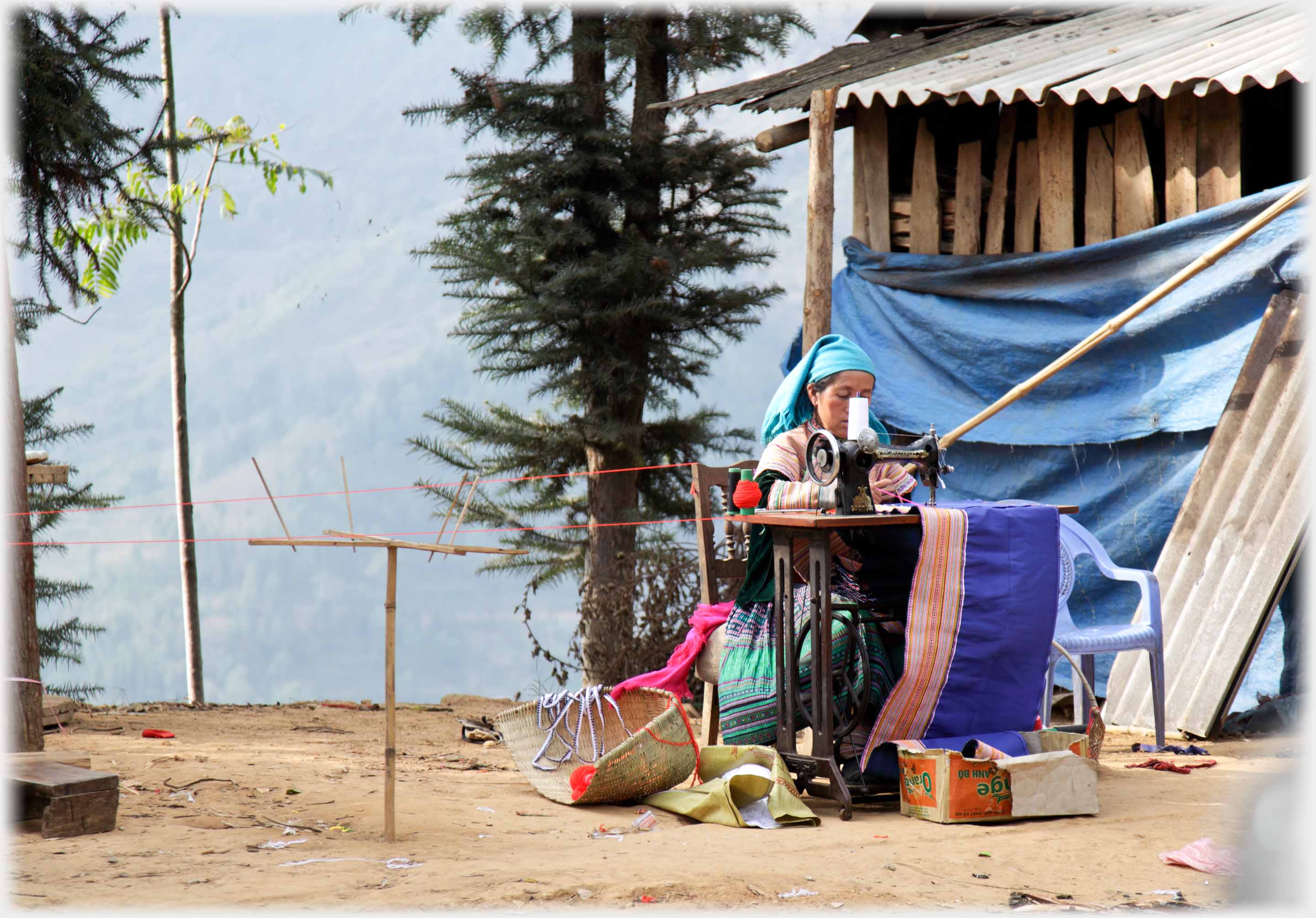 Woman sitting at machine with large cloth going through it, in front of shed, big valley behind.